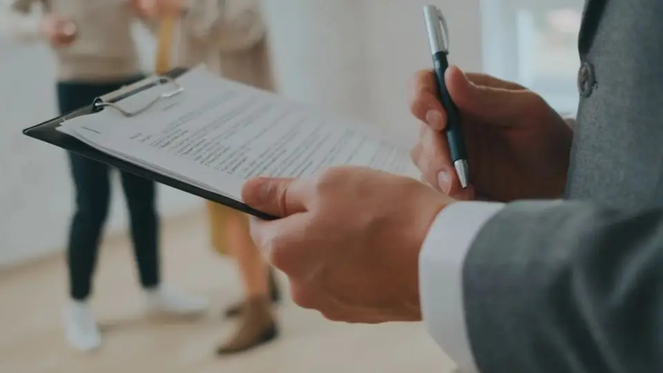 A clipboard and pen held in a bright empty room while two people walk through it
