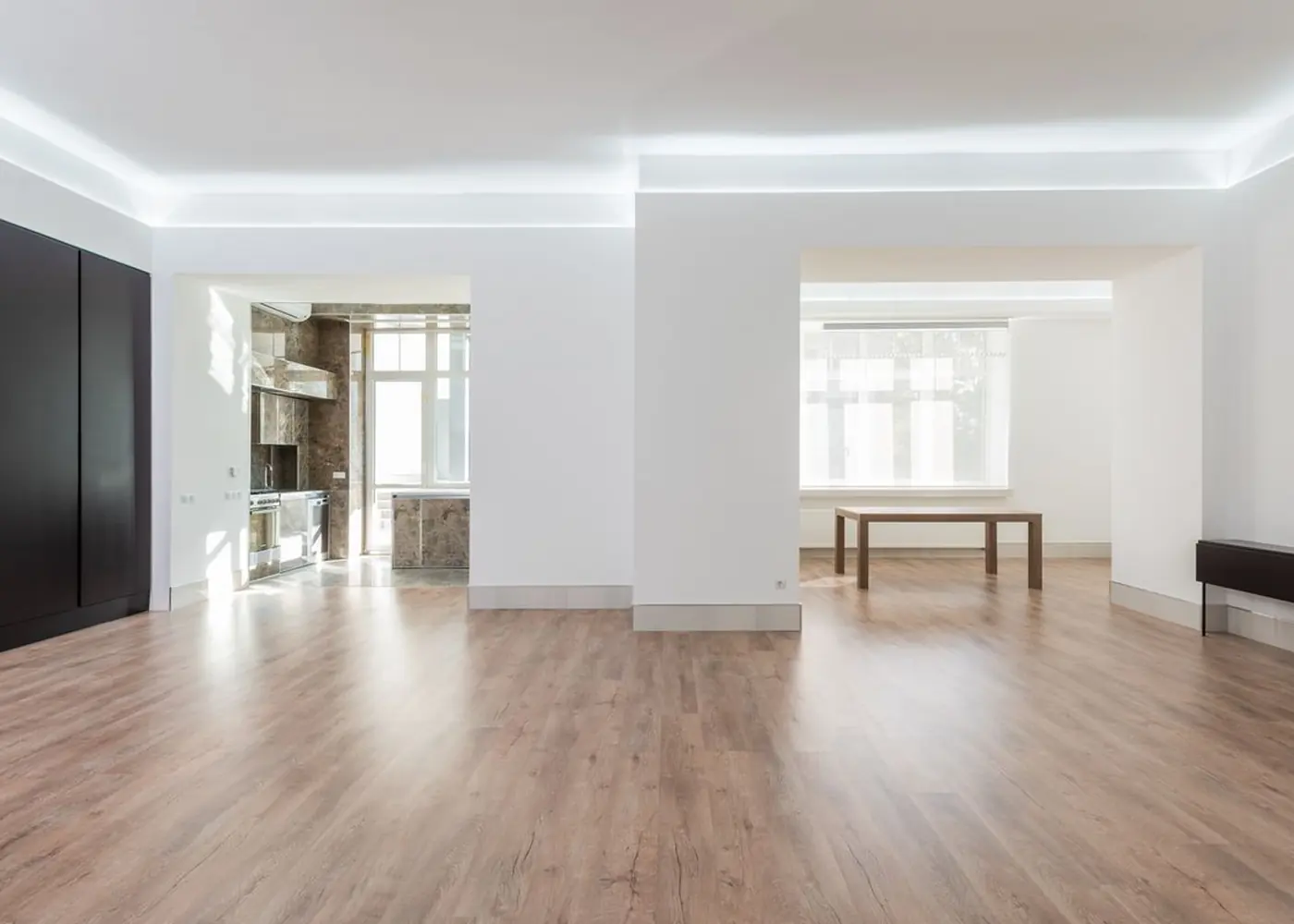 An empty unfurnished flat with pale walls, wooden flooring and a fitted kitchen visible through an open doorway