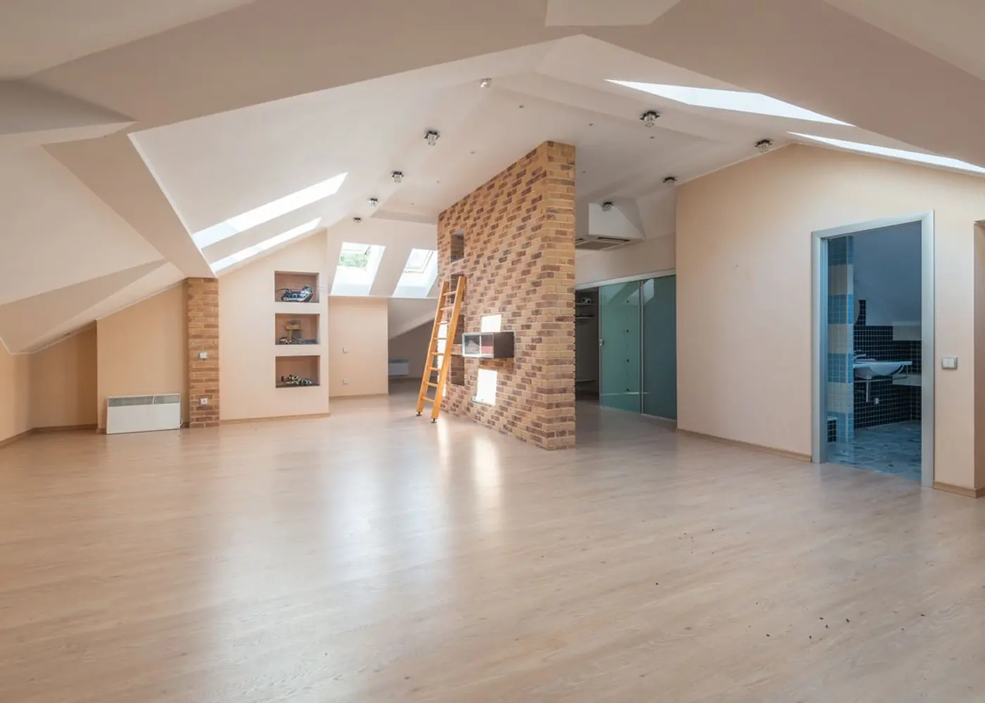 A bright open-plan apartment with skylights, exposed brick and pale timber floors, viewed from one end of the room